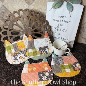 May include: Four quilted cat-shaped pot holders with patchwork patterns in shades of orange, yellow, green, and white. A white sign with the text "come together for food & memories" is in the background. A white teacup with a spoon is also visible.