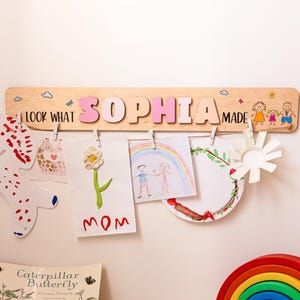 以下が含まれることがあります： ピンク色の文字で「SOPHIA」と書かれた木製のディスプレイボードには、子供たちの作品が飾られています。「LOOK WHAT MADE」という言葉と装飾的な絵が描かれています。虹色の木製おもちゃと本が入ったバスケットも表示されています。