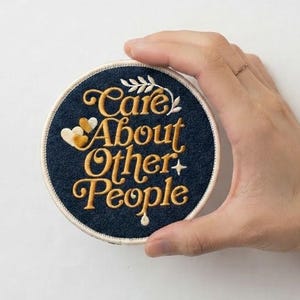 May include: A circular dark blue embroidered patch featuring the gold-colored text Care About Other People in a cursive font. The design includes small gold hearts, a leaf sprig, and a star accent, all bordered by a white stitched edge.