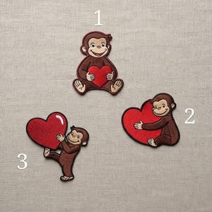 May include: Three embroidered patches featuring a brown monkey character holding a large red heart. Patch 1 shows the monkey sitting, patch 2 shows the monkey hugging a heart from the side, and patch 3 shows the monkey standing while holding a heart.