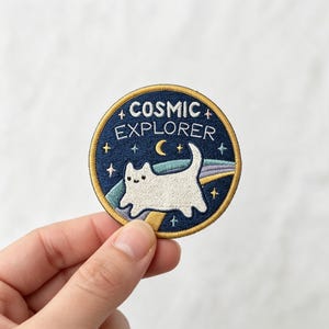 May include: A circular embroidered patch with a dark blue background featuring a white cat, a yellow crescent moon, and colorful stars. The text COSMIC EXPLORER is stitched in white across the top. The patch has a gold-colored border.