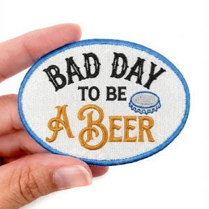 May include: An oval-shaped embroidered patch with a white background and a blue border. The text reads "BAD DAY TO BE A BEER" in black and gold lettering, accompanied by an embroidered graphic of a small blue and silver bottle cap.