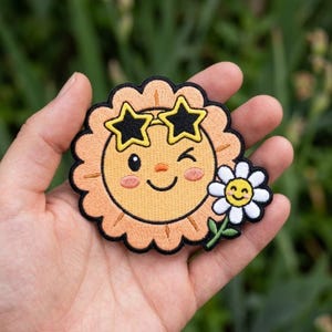 May include: An embroidered patch featuring a round, peach-colored sun character with a smiling face, winking eye, and black star-shaped sunglasses. A small white daisy with a yellow center is attached to the side of the sun character.