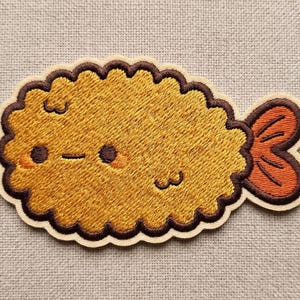 May include: Embroidered patch featuring a cute tempura shrimp design. The patch has a golden-yellow body with a brown scalloped edge and a small orange tail. The shrimp has a smiling face with rosy cheeks.