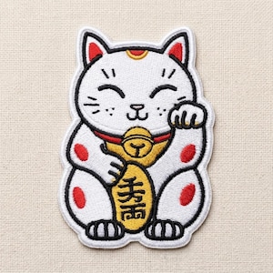 May include: A white embroidered patch shaped like a beckoning cat with black outlines. The cat features red ear accents, red spots on its body, a gold collar with a bell, and holds a gold oval coin with black Japanese kanji characters printed on it.