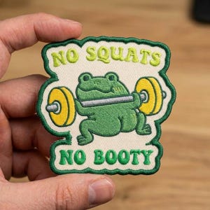 May include: An embroidered patch with a cream-colored background and a dark green border. It features a green frog lifting a barbell with yellow weights. The text NO SQUATS is embroidered in light green above and NO BOOTY in green below.