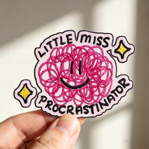 May include: A light pink embroidered patch featuring a dark pink scribbled circle with a black smiley face. The text LITTLE MISS PROCRASTINATOR is stitched in black around the center. Two small yellow four-pointed stars are on the sides.