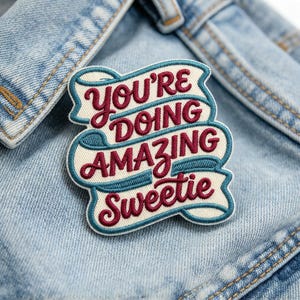 May include: An embroidered patch featuring the text "YOU'RE DOING AMAZING Sweetie" in dark red lettering. The words are set against a cream-colored background shaped like a ribbon, with a teal blue border outlining the entire design.