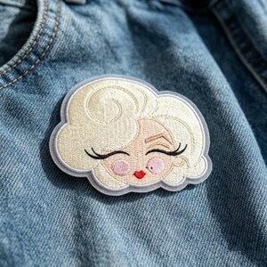 May include: An embroidered patch featuring a stylized face with platinum blonde hair, long black eyelashes, pink cheeks, and bright red lips. The patch has a light gray border and is attached to a blue denim fabric background.