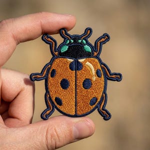 May include: An embroidered patch shaped like a beetle with an orange body, dark blue spots, and a dark blue border. The head is black with green accents, and the legs are orange with dark blue outlines. The design features a symmetrical insect.