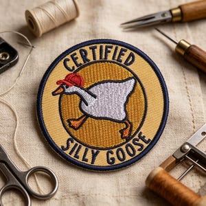 May include: A circular embroidered patch featuring a white goose wearing a red baseball cap. The background is yellow with a dark blue border. The text "CERTIFIED SILLY GOOSE" is embroidered in dark blue lettering along the top and bottom edges.
