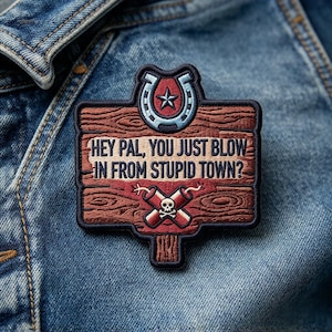 May include: A rectangular embroidered patch shaped like a wooden sign with a dark blue border. It features a light blue horseshoe with a star, the text HEY PAL, YOU JUST BLOW IN FROM STUPID TOWN?, and two crossed red dynamite sticks with a skull.