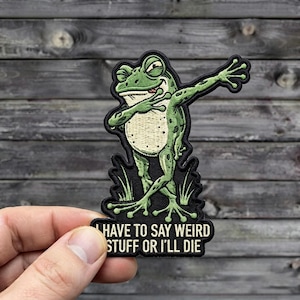 May include: An embroidered patch featuring a green frog performing a dab pose. The frog has a cream-colored belly and is surrounded by small grass tufts. Below the frog, the text reads: I HAVE TO SAY WEIRD STUFF OR I'LL DIE in white letters.