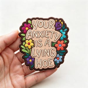 May include: An embroidered patch with a light beige background and a dark brown border. The text reads YOUR ANXIETY IS A LYING HOE in dark brown letters. The design is surrounded by colorful embroidered flowers in pink, yellow, purple, orange, and blue.