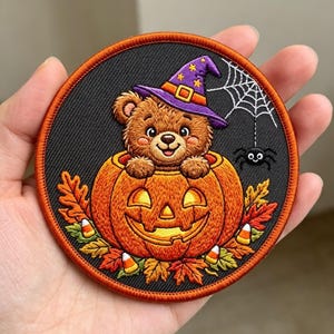 May include: A circular embroidered patch featuring a brown teddy bear wearing a purple witch hat, sitting inside a carved orange jack-o'-lantern pumpkin. The design includes autumn leaves, candy corn, a white spider web, and a small black spider.