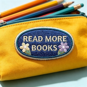 May include: A yellow pencil case featuring an oval embroidered patch with a dark blue background. The patch displays the white text READ MORE BOOKS, flanked by a light yellow flower on the left and a light purple flower on the right side.