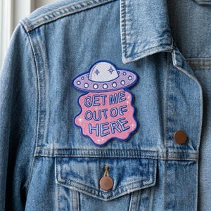 May include: An embroidered patch attached to a blue denim jacket. The patch features a purple and white UFO hovering above a pink, cloud-shaped background with the blue text GET ME OUT OF HERE and small white star accents embroidered on the surface.