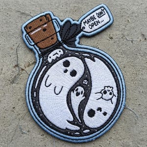 May include: An embroidered patch shaped like a glass bottle with a brown cork stopper. Inside the bottle, white ghosts form a yin-yang pattern against a black background. A small tag attached to the neck reads MAYBE DON'T OPEN in black text.