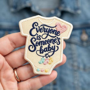 May include: A cream-colored embroidered patch shaped like a baby onesie. It features dark blue cursive text reading Everyone is Someone's baby, a small pink heart, three tiny hanging baby socks in pastel colors, and three small buttons.