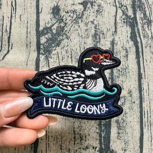 May include: An embroidered patch featuring a black and white loon wearing red heart-shaped sunglasses. The bird floats on stylized teal water waves above a dark blue banner with the white embroidered text LITTLE LOONY in a bold font.