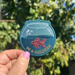 May include: A circular embroidered patch featuring a teal fishbowl with a pink goldfish swimming inside. The text BE A GOLDFISH is stitched in gold thread along the bottom curve of the bowl, which has a dark teal water line and white highlights.
