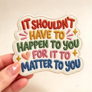 May include: An embroidered patch with a cream-colored background and scalloped edges. The text reads IT SHOULDN'T HAVE TO HAPPEN TO YOU FOR IT TO MATTER TO YOU in red, yellow, green, pink, and blue thread, accented by small stars and a heart.