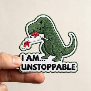 May include: An embroidered patch featuring a green Tyrannosaurus Rex holding two small red dinosaur skulls on sticks. The patch has a white background with the black text "I AM... UNSTOPPABLE" and a dark green border, creating a humorous design.