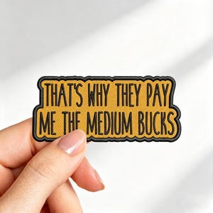May include: A rectangular embroidered patch with a scalloped black border and a golden yellow background. The text "THAT'S WHY THEY PAY ME THE MEDIUM BUCKS" is stitched in black, sans-serif capital letters across the center of the patch.