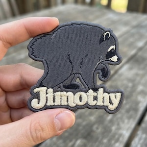 May include: An embroidered patch featuring a dark gray raccoon in a walking pose with a white face mask and ears. Below the raccoon, the name Jimothy is stitched in large, cream-colored, rounded letters with a dark gray outline on a gray background.