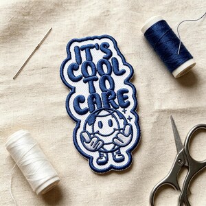 May include: An embroidered patch featuring the text IT'S COOL TO CARE in dark blue thread on a white background. Below the text is a cartoon globe character with a smiling face, arms, and legs. The patch is surrounded by a dark blue border.