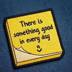 May include: Yellow embroidered patch with the text "There is something good in every day" and a smiling face. The patch has a black border and a small corner that is peeled back. The patch is on a blue denim background.