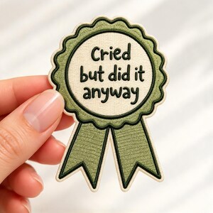 May include: A circular embroidered patch shaped like a ribbon award with a scalloped edge. The center is cream-colored with the black text Cried but did it anyway. The border and two hanging ribbons are olive green with dark green outlines.