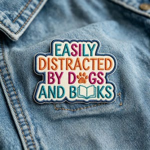 May include: An embroidered patch with a cream-colored background and dark blue border. The text reads EASILY DISTRACTED BY DOGS AND BOOKS, with a brown paw print replacing the O in DOGS and an open book icon replacing the OO in BOOKS.