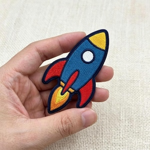 May include: An embroidered rocket ship patch featuring a blue body, a yellow nose cone, a white circular window, and red side fins. The rocket is depicted with a yellow and orange flame emerging from the bottom, all outlined in dark navy blue thread.