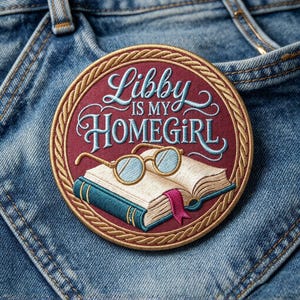 May include: A circular embroidered patch with a deep red background and a gold rope-style border. The text reads Libby IS MY HOMEGIRL in light blue script and block letters. An open book with a pink bookmark and gold-rimmed glasses are centered.
