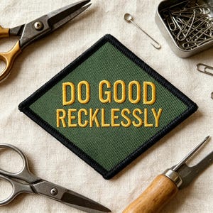 May include: A diamond-shaped embroidered patch with a dark olive green background and a thick black border. The text DO GOOD RECKLESSLY is stitched in bold yellow capital letters across the center of the patch, featuring a clean sans-serif font.