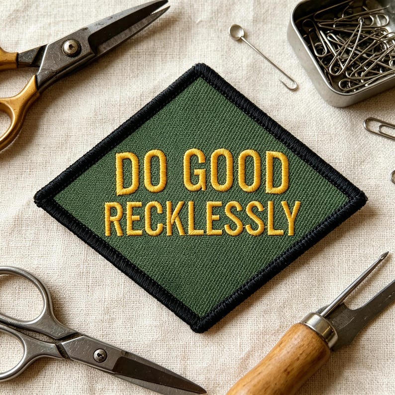 May include: A diamond-shaped embroidered patch with a dark olive green background and a thick black border. The text DO GOOD RECKLESSLY is stitched in bold yellow capital letters across the center of the patch, featuring a clean sans-serif font.