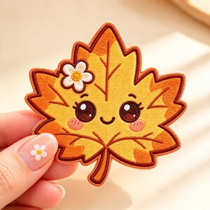 May include: An embroidered patch shaped like a maple leaf in shades of yellow and orange. The leaf features a smiling face with large dark eyes, pink cheeks, and a small white flower detail near the top edge, outlined in dark brown thread.
