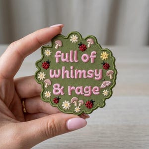 May include: An olive green embroidered patch with a scalloped edge featuring the pink text full of whimsy & rage. The design includes small white flowers, red ladybugs, light pink mushrooms, and a small moth scattered across the green surface.