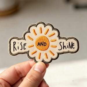 May include: An embroidered patch featuring a central orange sun with yellow rays on a cream-colored background. The text "RISE AND SHINE" is stitched in black thread across the design, which is outlined with a dark brown border.