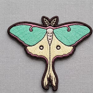 May include: An embroidered patch shaped like a moth with mint green upper wings, cream lower wings, and a dark brown border. The moth features a cream-colored body, small antennae, and pink accents along the wing edges, all stitched with thread.