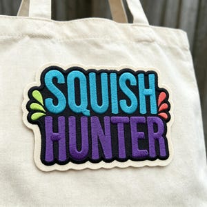 May include: A cream-colored canvas tote bag featuring a large embroidered patch. The patch displays the text SQUISH HUNTER in bright blue and purple letters against a black background, accented by small green and coral-colored leaf shapes.