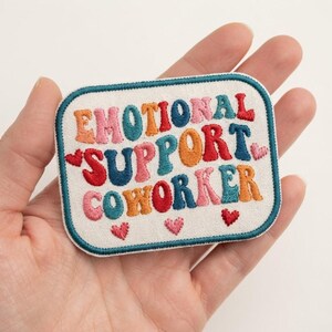 May include: A rectangular embroidered patch with rounded corners on a cream-colored fabric base. The text reads EMOTIONAL SUPPORT COWORKER in wavy, multi-colored letters including red, blue, pink, and orange. Three small pink hearts are below.