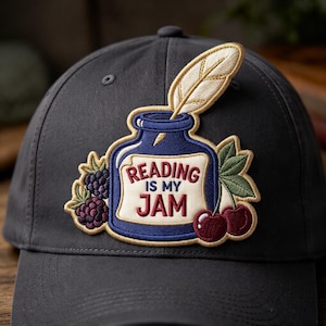May include: A dark gray baseball cap featuring an embroidered patch of a blue inkwell with a quill pen, blackberries, and cherries. The patch includes the text "READING IS MY JAM" in red lettering on a cream-colored label on the inkwell.
