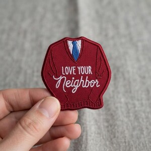 May include: A maroon embroidered patch shaped like a V-neck sweater with a white shirt collar and a blue necktie. The text "LOVE YOUR Neighbor" is stitched in white thread across the center of the sweater design with a ribbed hem detail.