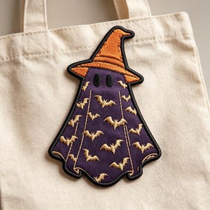 May include: A beige canvas tote bag featuring an embroidered patch of a ghost wearing an orange witch hat. The ghost is purple with a repeating pattern of small gold bats and has two black oval eyes, all outlined in a thick black border.