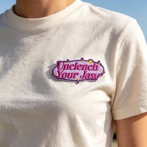 May include: A cream-colored short-sleeve t-shirt featuring an embroidered patch on the chest. The patch is a purple oval shape with a dark purple border, containing the text "Unclench Your Jaw" in pink letters, accented by small yellow stars.