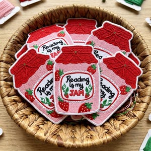 May include: A collection of embroidered patches shaped like strawberry jam jars, each featuring a red lid, a light pink jar body, and the text Reading is my JAM in black lettering, surrounded by small embroidered strawberry illustrations.