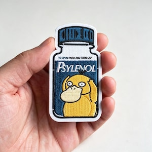 May include: An embroidered patch shaped like a medicine bottle with a dark blue cap and label. The label features the text "TO OPEN: PUSH AND TURN CAP" and "Psylenol" in white, with a yellow duck-like creature graphic centered below the text.