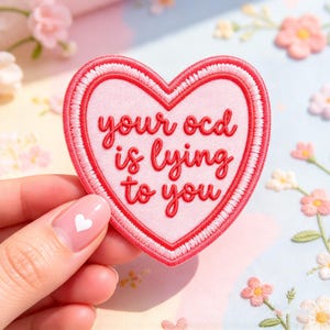 May include: A heart-shaped embroidered patch with a light pink center and a red border. The text "your ocd is lying to you" is stitched in red cursive lettering across the center of the heart, featuring a white inner border detail.